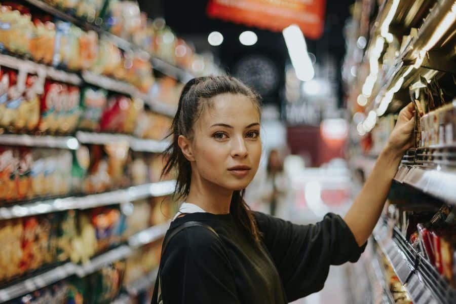 Young woman reaches toward a grocery shelf in a supermarket aisle and looks back toward the camera, with rows of packaged products blurred behind her. It suits retailer tricks content about how product placement can influence what shoppers notice and choose.