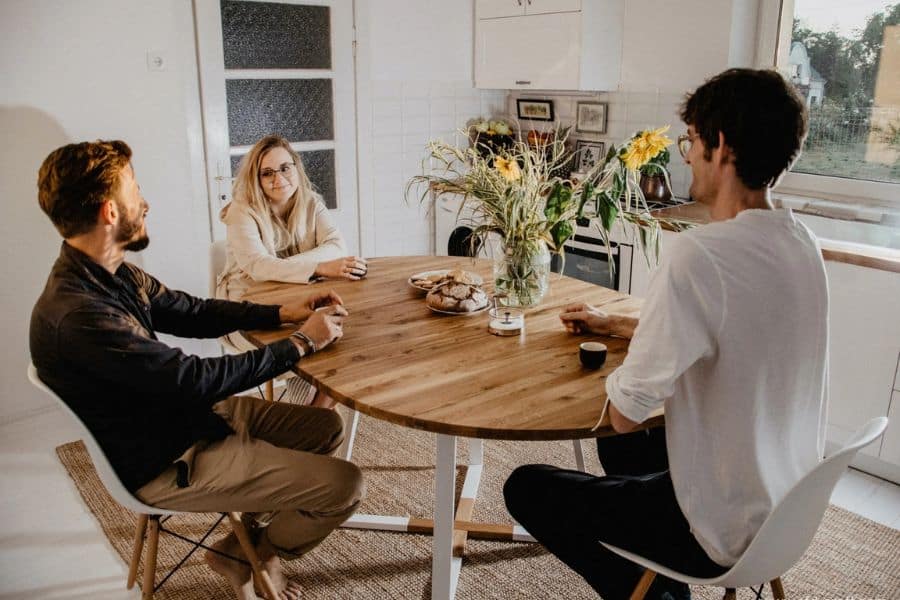 Three people sit around a wooden kitchen table with coffee cups, flowers, and pastries in a bright cozy home. This warm gathering reflects Scandinavian lifestyle habits focused on connection, comfort, and relaxed time together indoors.
