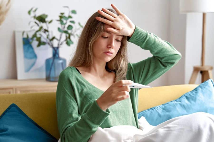 Young woman sits on a couch under a blanket while checking a thermometer and touching her forehead. The photo conveys fever or fatigue during illness.
