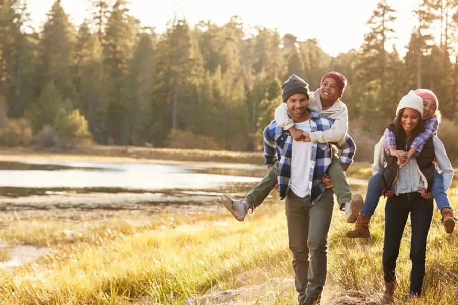 Two adults carry children on their backs while walking beside a lake with tall trees glowing in soft golden light. The relaxed family outing fits Scandinavian lifestyle habits that value outdoor time and togetherness across seasons.