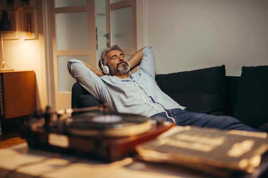 Middle aged man reclining on a dark sofa with headphones on while listening quietly in a warmly lit living room. A record player in the foreground suggests relaxing audio used to unwind after lack of sleep.