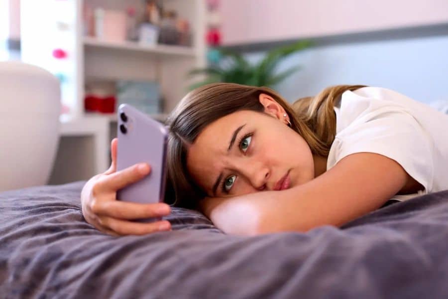 Young woman lying on a bed and staring at her phone with a tired and distant expression. The scene suggests one of the destructive habits of doomscrolling or withdrawing into screen time when feeling low.