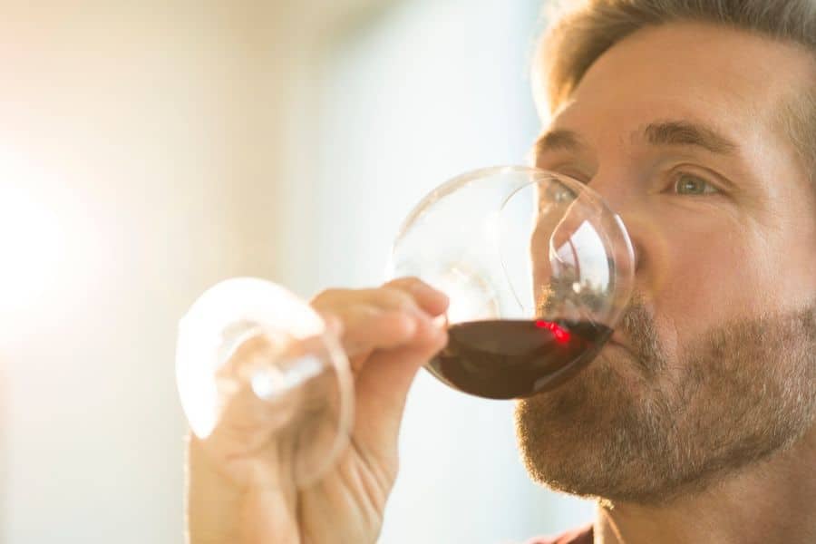 A man drinks red wine from a glass while looking off to the side in soft natural light. The close up focuses on the act of sipping, suggesting relaxation or lifestyle habits involving alcohol.