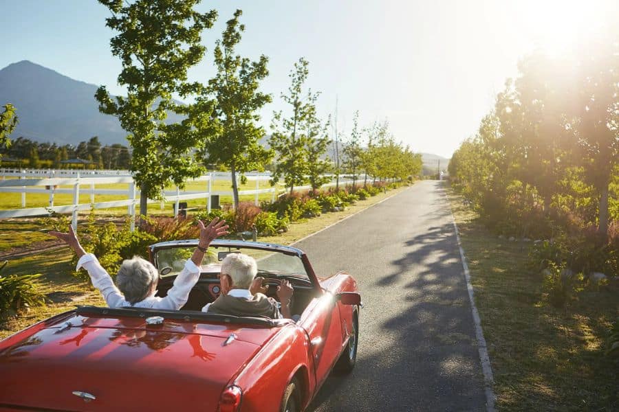 Older couple riding in a red convertible down a sunlit country road lined with trees and white fences. One passenger raises both arms in celebration to suggest the freedom that thoughtful financial strategies can support later in life.
