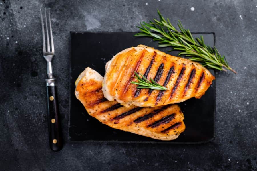 Grilled chicken breasts with dark grill marks rest on a black plate with sprigs of rosemary on a dark stone surface. A fork beside the plate frames the dish as a prepared high protein meal.