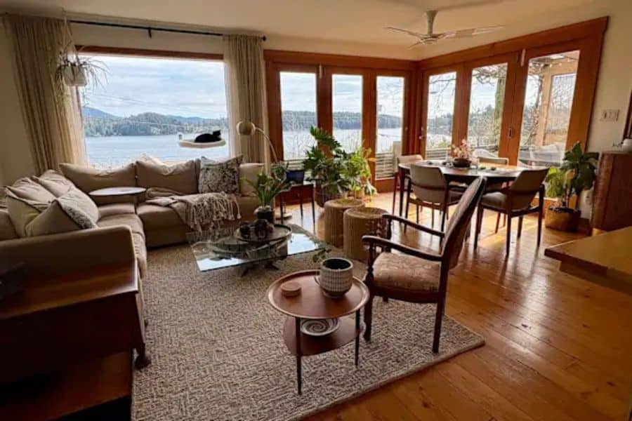Warm, cozy living room with a beige sectional sofa, woven rug, wood floors, indoor plants, and a dining area beside large windows overlooking a calm lake. The space highlights a comfortable home stay option for travelers trying to see the world on a budget.