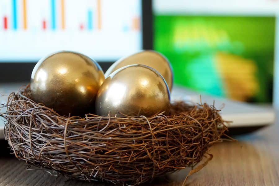 Three metallic gold eggs sit in a twig nest on a desk with blurred charts glowing in the background. The visual uses the golden eggs as a metaphor for protected savings and future wealth.