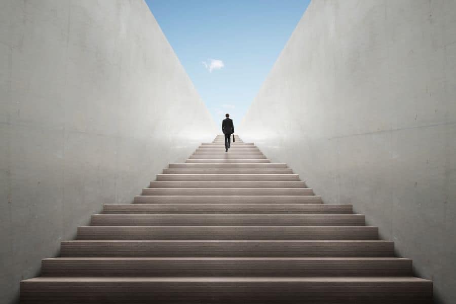 A lone person in a dark suit walks up a long, wide staircase between tall concrete walls toward an open blue sky. The upward path visually represents progression and life journey in a post about how long you're going to live.