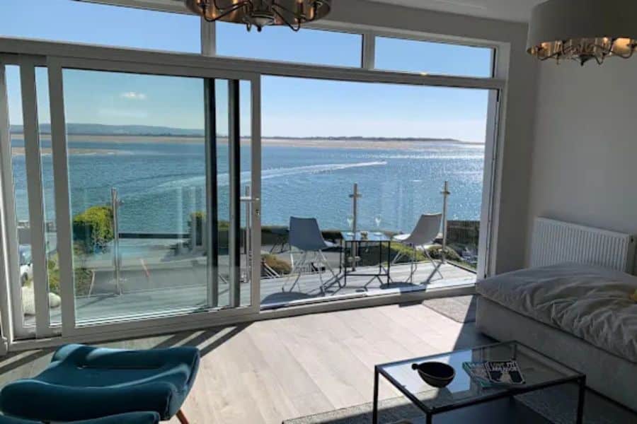 Bright modern room with a bed, glass coffee table, blue chair, and wide sliding doors opening to a patio with two chairs facing a sparkling ocean view. The scene shows how see the world on a budget can include simple accommodations with memorable waterfront views.
