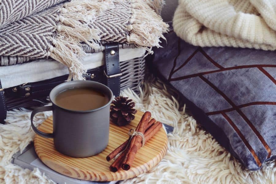 A mug of tea sits on a wooden tray beside cinnamon sticks, knit blankets, and textured pillows on a soft rug. The cozy setup reflects Scandinavian lifestyle habits built around warmth, rest, and simple comforting details at home.