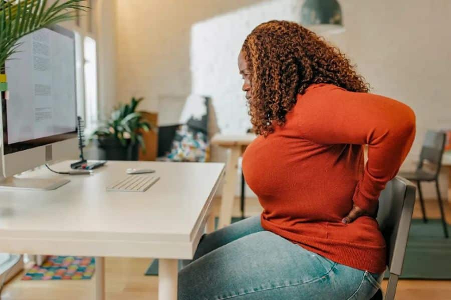 A woman sitting at a desk leans forward while holding her lower back in pain. This posture reflects muscle discomfort or tension that can be connected to being low in magnesium.