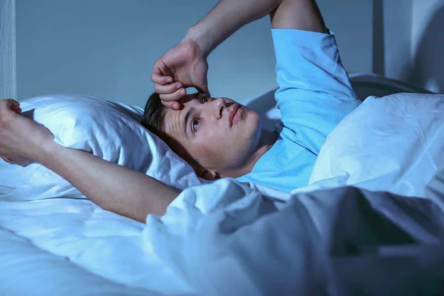 A man lies awake in bed at night with his hand on his forehead, staring upward. The restless scene illustrates sleep issues commonly linked to low in magnesium levels.