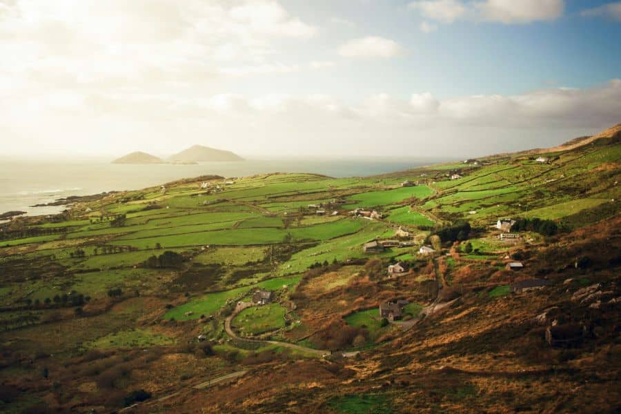 A wide view of green coastal farmland with scattered houses winding roads and the ocean in the distance under soft sunlight. The landscape shows a serene countryside scene that appeals to Americans seeking natural beauty abroad.