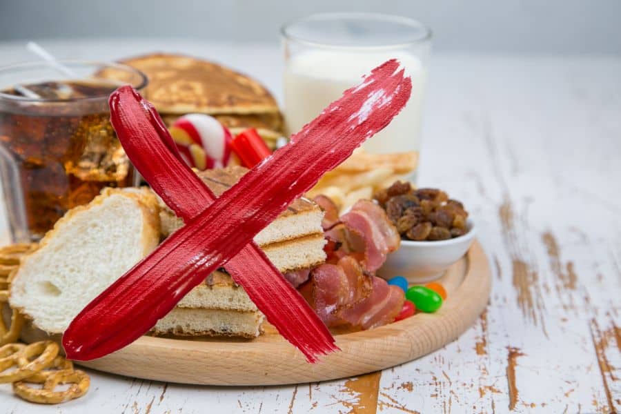 Assorted processed foods and sugary drinks on a wooden board are covered with a large red painted X. The image signals foods to avoid when managing chronic inflammation.