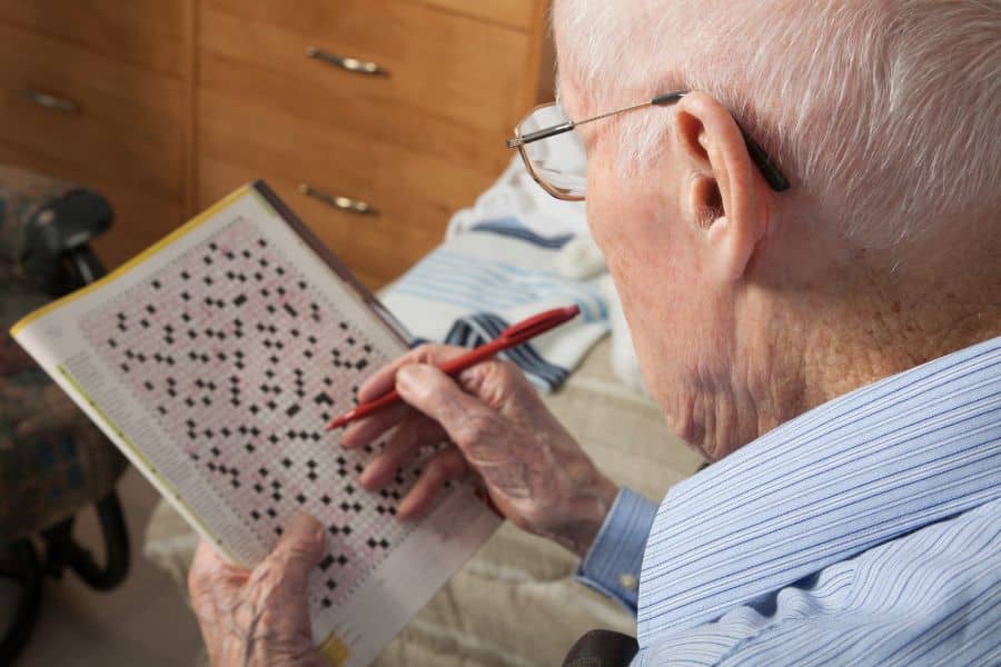 Older man wearing glasses sits indoors and works on a puzzle book with a red pen in hand. This fits the theme of living a longer life by showing a quiet activity that suggests focus and mental engagement.