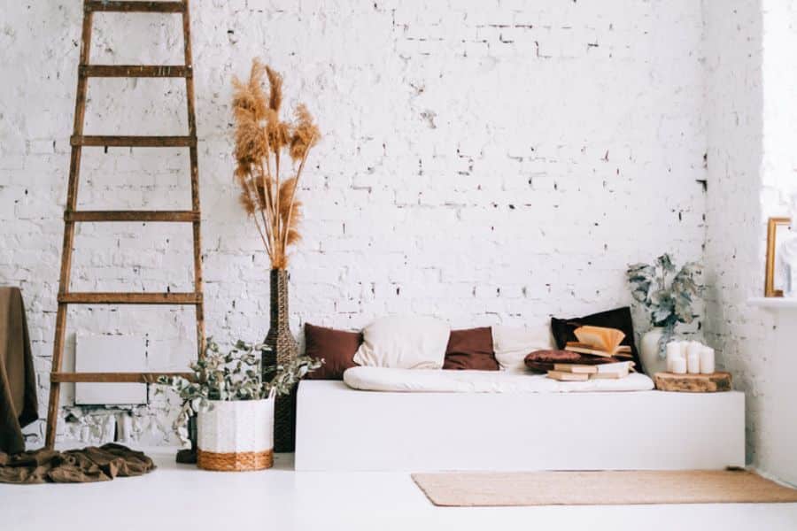 A minimalist room with a white brick wall features a low daybed, neutral pillows, stacked books, candles, plants, and a wooden ladder. This calm interior shows Scandinavian lifestyle habits through uncluttered design and cozy natural textures.