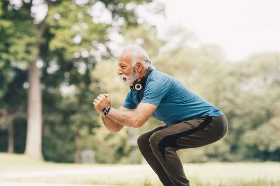 Older man exercising outdoors in a park while holding a deep squat, with headphones around his neck and trees blurred in the background. The photo highlights strength and mobility during older adulthood.
