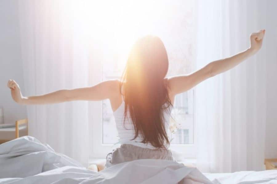 Woman sitting up in bed and stretching toward a bright window with sunlight shining around her hair. The soft white room suggests a fresh start to the morning after lack of sleep.