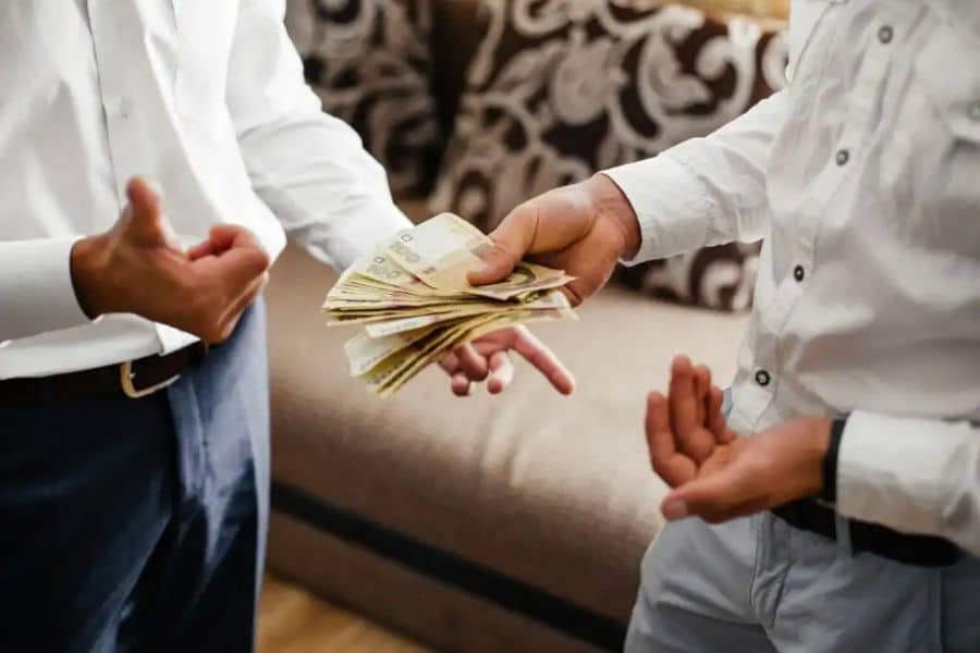 One person hands a stack of cash to another in a living room while both stand facing each other. This photo represents a direct cash loan or private payment exchange.