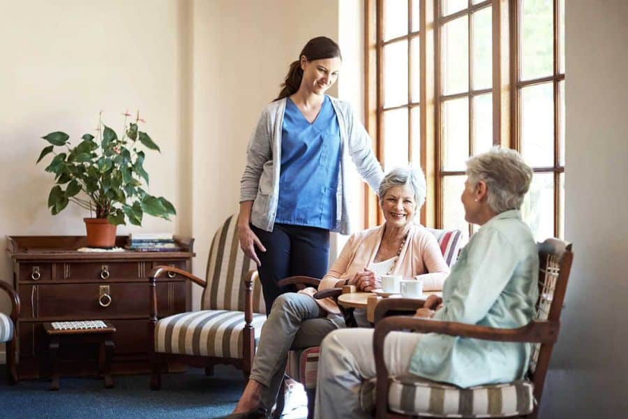 Caregiver standing beside two older women seated and talking in a bright living room. The photo represents supportive senior care and the role financial strategies can play in planning for daily assistance.