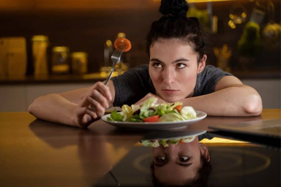A woman sits at a table staring at a plate of salad while holding a fork with little interest. Her lack of appetite and disengaged expression can relate to feeling low in magnesium.