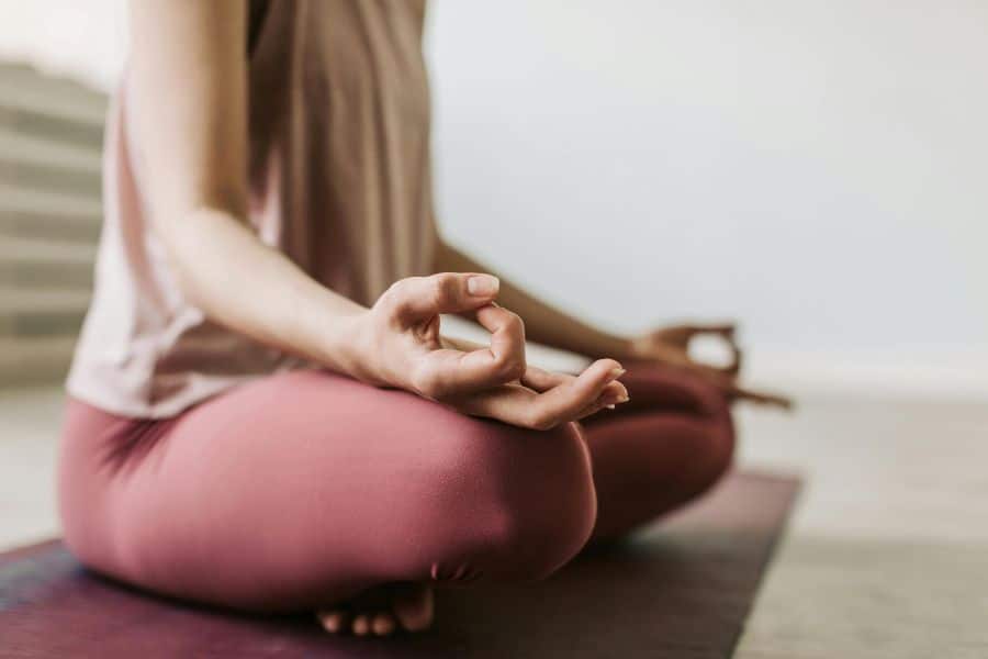 Person seated cross legged on a yoga mat with one hand resting on the knee in a meditation pose. The soft indoor scene suggests stress reduction practices that may support chronic inflammation management.