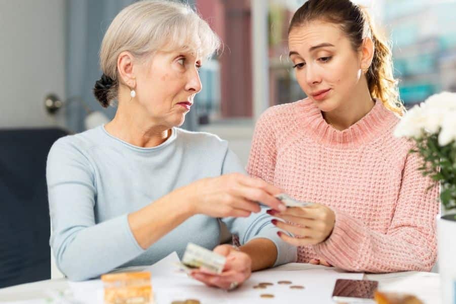 An older woman hands cash across a table to a younger woman while coins bills and a wallet sit nearby. The close interaction highlights direct financial support between generations.