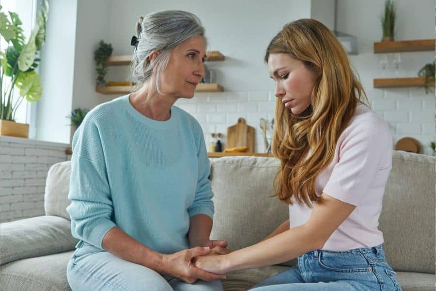 An older woman and a younger woman sit on a couch holding hands in a quiet supportive moment. Their expressions suggest an emotional conversation about stress hardship or family responsibilities.