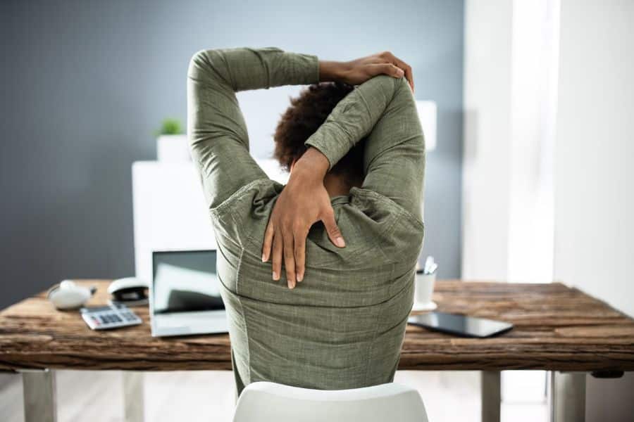 Person seen from behind sits at a desk and stretches one arm overhead while reaching the other hand down the back. A laptop, calculator, and tablet sit on the desk, showing a quick seated stretch during the workday.