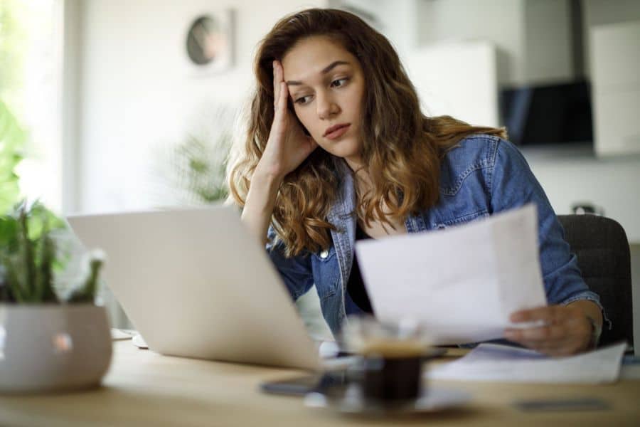 A young woman sits at a table with a laptop and paperwork, resting her head on one hand as she looks worried. This fits retirement savings content about financial stress, budgeting challenges, or feeling behind on long term goals.