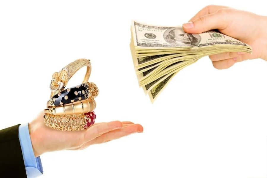 Hand holding several gold bracelets reaches toward another hand offering a stack of dollar bills on a white background. The image suggests exchanging jewelry for fast cash or a collateral based loan.