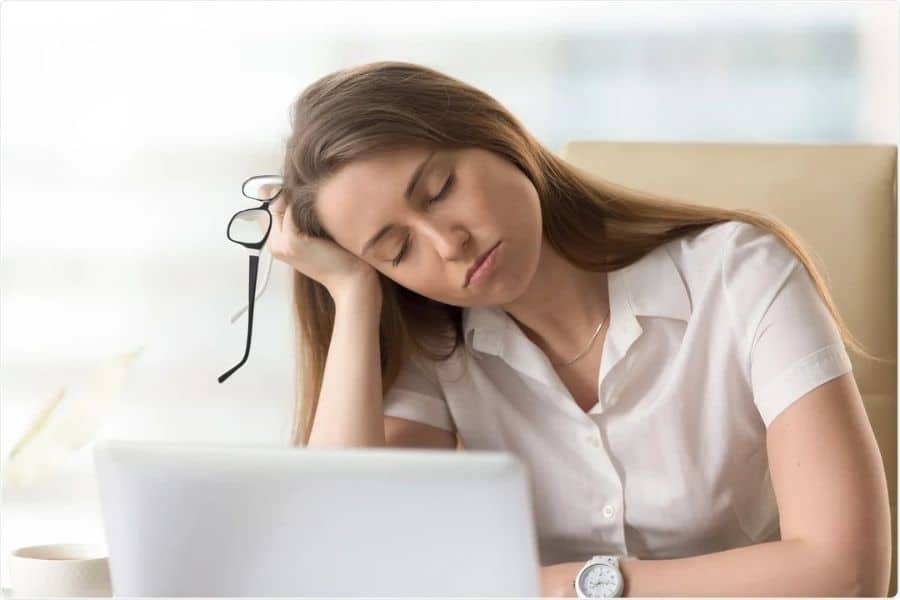 Tired office worker leans on one hand at a desk while holding eyeglasses beside an open laptop. The scene represents exhaustion or low energy that could relate to early stages of cancer.