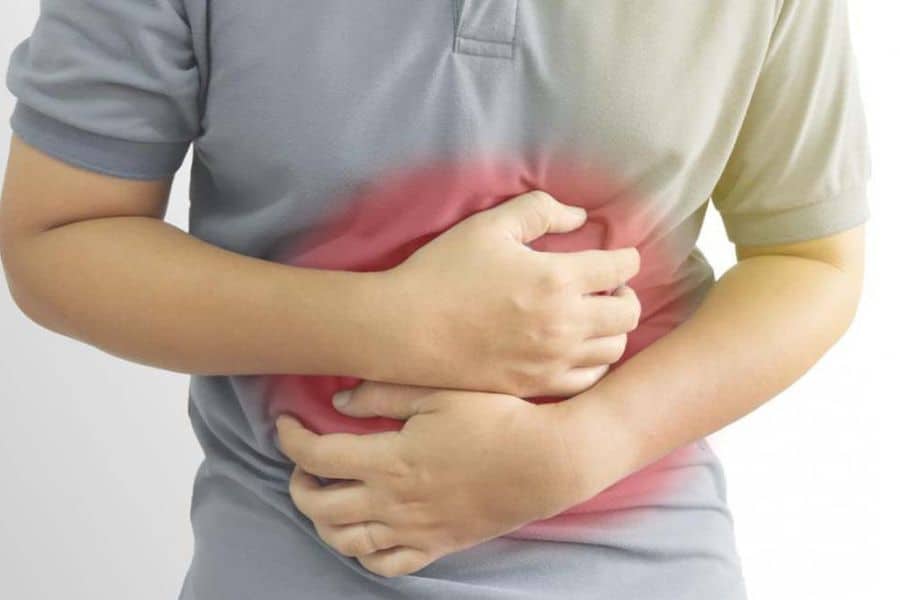 Person in a gray shirt clutches their abdomen with both hands while a red overlay marks the painful area. The photo illustrates sharp stomach or digestive pain.