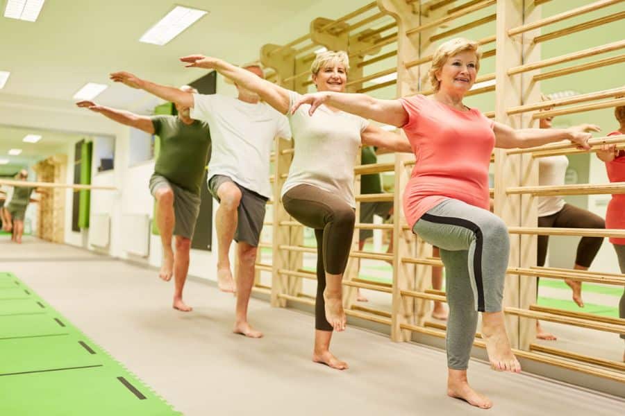 Group of older adults balances on one leg with arms extended during an exercise class beside wooden wall bars. The scene suggests guided movement practice that can improve balance, mobility, and posture habits.