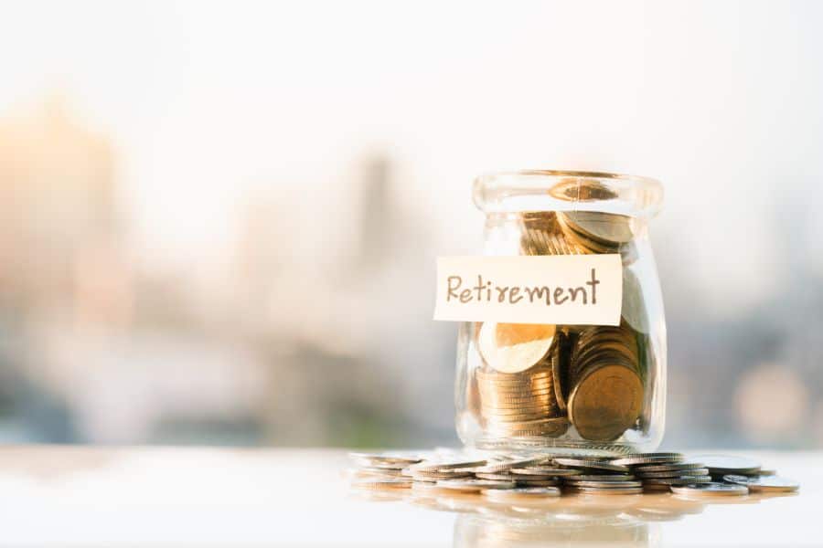 Glass jar filled with coins labeled "Retirement" sits on a table with more coins scattered around suggesting long term savings and paying less tax through planning. Soft blurred city background reinforces a financial future focus.