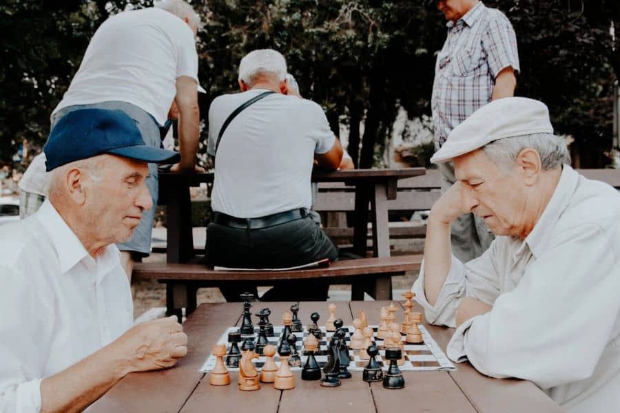 Two older men sit across from each other playing chess at a picnic table in a park while several other seniors gather behind them. The scene emphasizes social connection and mental activity in later life.