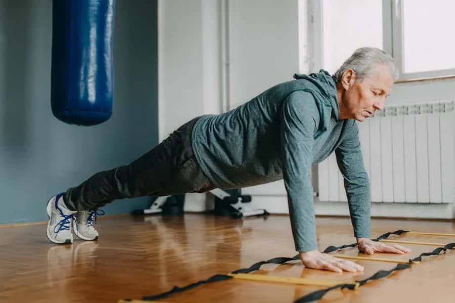 Older man holding a plank position on a gym floor beside an agility ladder, with a punching bag hanging in the background. The photo shows upper body and core exercise for healthy aging.