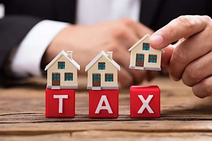 Small house models placed on blocks spelling "TAX" with a hand holding another house symbolizing property taxes and ways of paying less tax on real estate. Wooden table background emphasizes financial planning concepts.