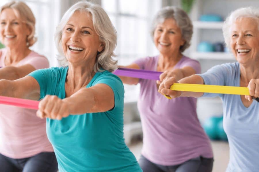 Four older women smile while holding colorful resistance bands stretched forward during a group workout class. This demonstrates joint strengthening exercises that improve upper body strength and stability in a supportive group setting.
