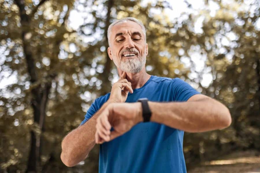Gray haired man in a blue shirt smiles while checking his smartwatch outdoors with trees softly blurred behind him. The image suggests tracking fitness or health habits during older adulthood.