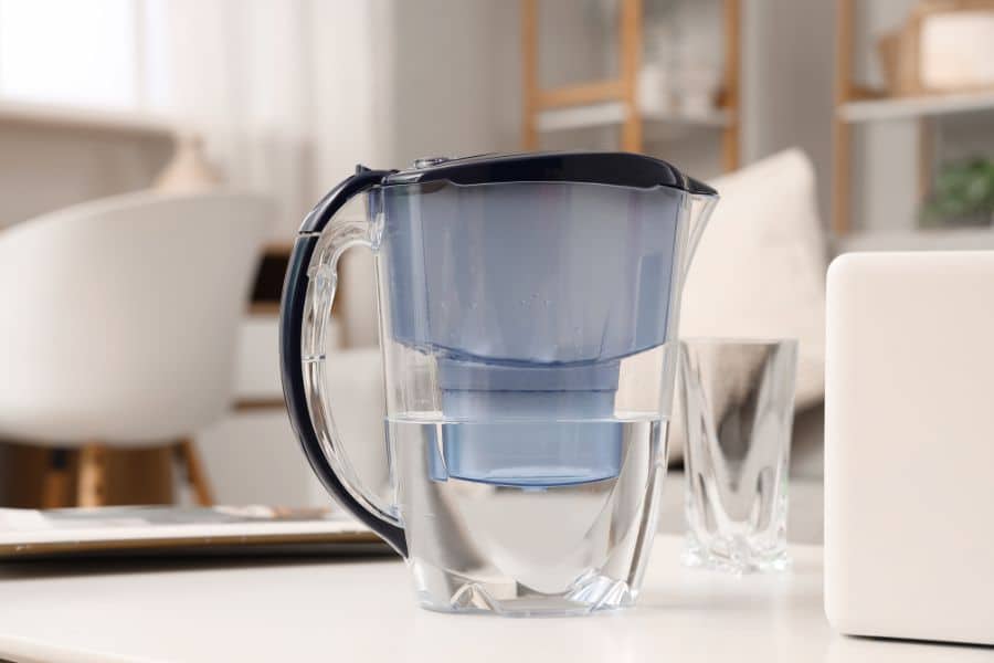 Clear water filter pitcher on a tabletop beside an empty drinking glass in a bright home setting. The image reinforces clean drinking water as part of healthy daily habits.