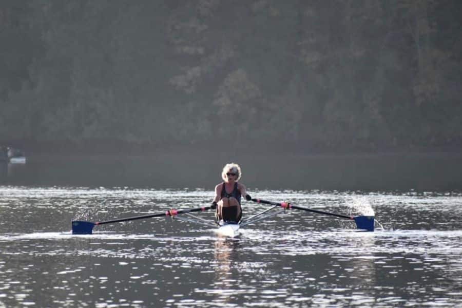 Older adult rows a narrow sculling boat across a calm lake with tree covered shoreline in the distance. The image shows a low impact outdoor workout that can help build strength around the joints.