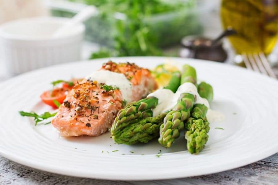 White dinner plate with a cooked salmon fillet, several asparagus spears, and a light creamy sauce, with blurred herbs and oil in the background. This plated meal fits content about foods that stop memory loss by showing fish and vegetables often linked with brain healthy eating.