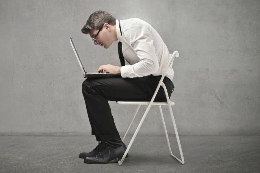 Man in office clothes sits on a folding chair and hunches far forward toward a laptop with his neck craned and shoulders rounded. The photo demonstrates a strained seated position often caused by unhealthy posture habits.
