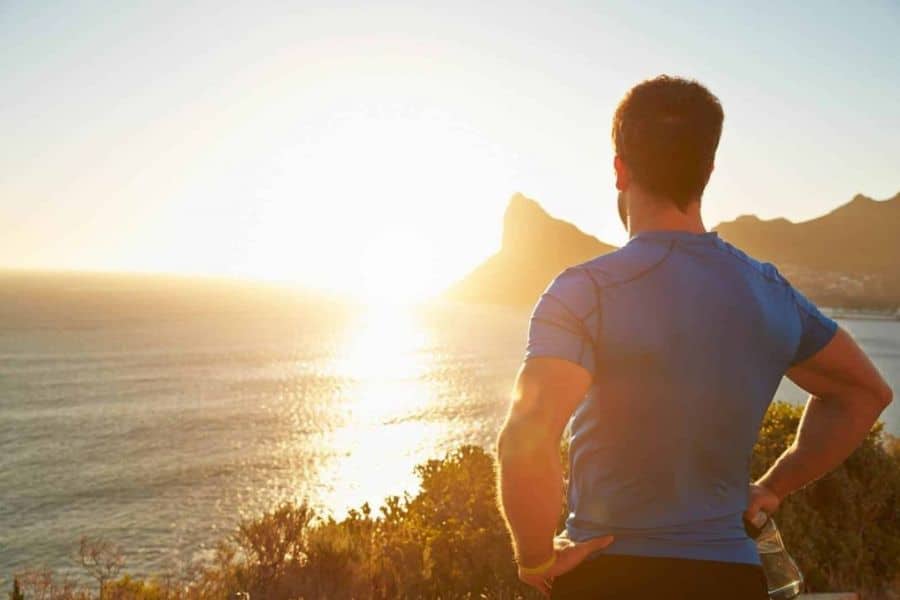 Man in a blue athletic shirt stands on a coastal overlook at sunrise with his hands on his hips and a water bottle at his side. The ocean glows in the morning light with mountains rising in the distance.