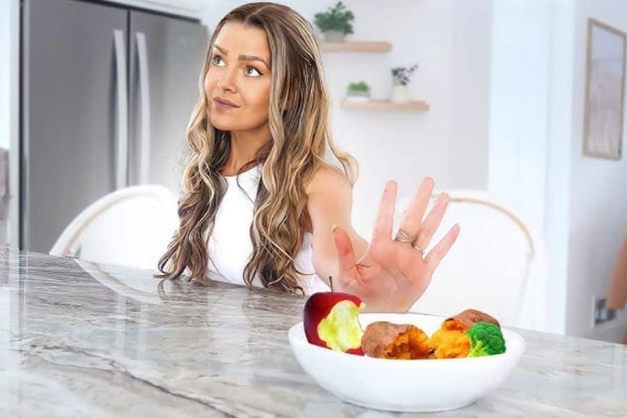 Woman sitting at a kitchen counter raises her hand to refuse a bowl of food containing an apple core broccoli and roasted vegetables. She looks off to the side as if making a careful food or health choice.