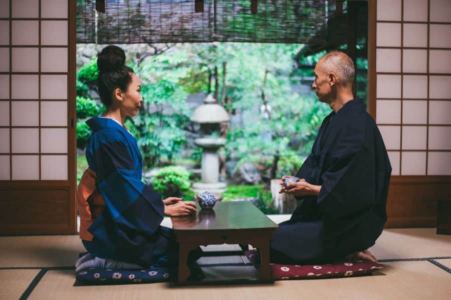 Two adults in traditional Japanese clothing kneel across from each other at a low table in a tatami room. A lush green garden fills the open view behind them creating a calm reflective setting.