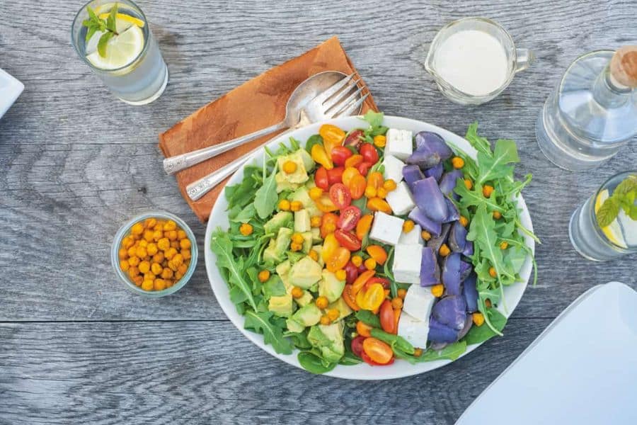 Overhead view of a colorful salad on a wooden table with avocado cherry tomatoes leafy greens white cheese cubes and purple vegetables. Glasses of water with lemon and a small bowl of crunchy snacks surround the plate suggesting a fresh balanced meal.