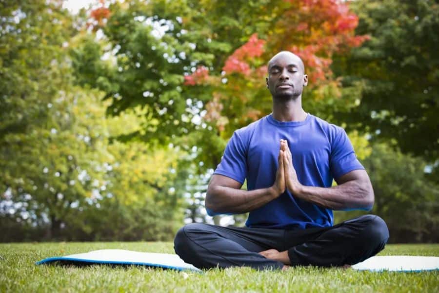 Man sits cross legged on a yoga mat in a grassy park with his eyes closed and hands pressed together in front of his chest. Trees with green and red leaves behind him create a peaceful scene that suits a Centenarian Secrets article about stress relief and wellbeing.