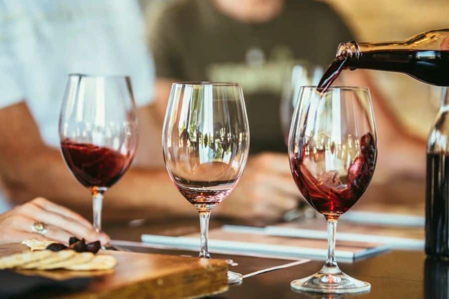 Red wine is being poured into a glass at a table set with other wine glasses and a small board of crackers and olives. Blurred people in the background suggest a relaxed social gathering.